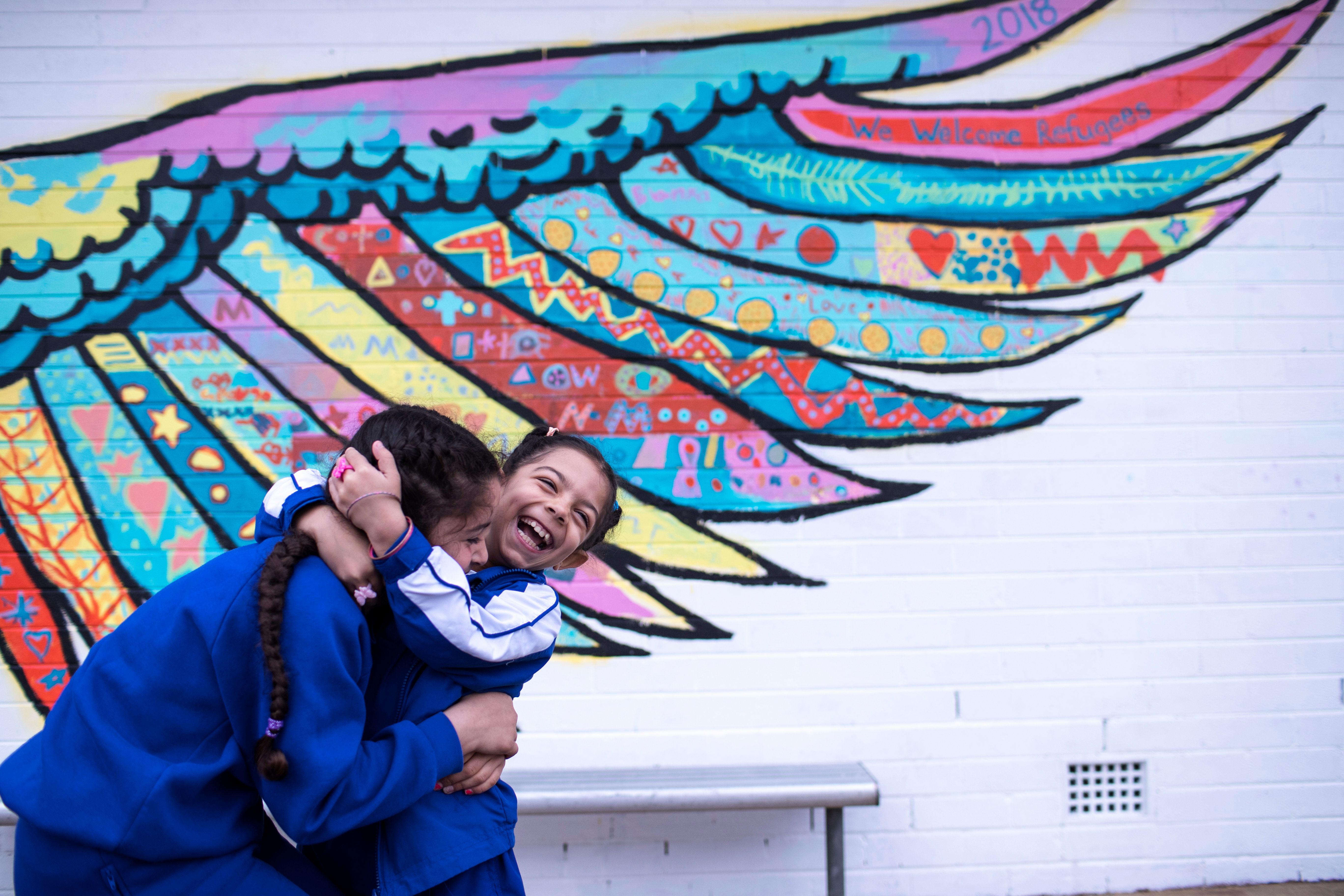 Two children hugging and laughing in front of a mural of wings painted on a wall 