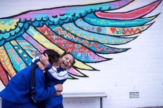 Two children hugging and laughing in front of a mural of wings painted on a wall