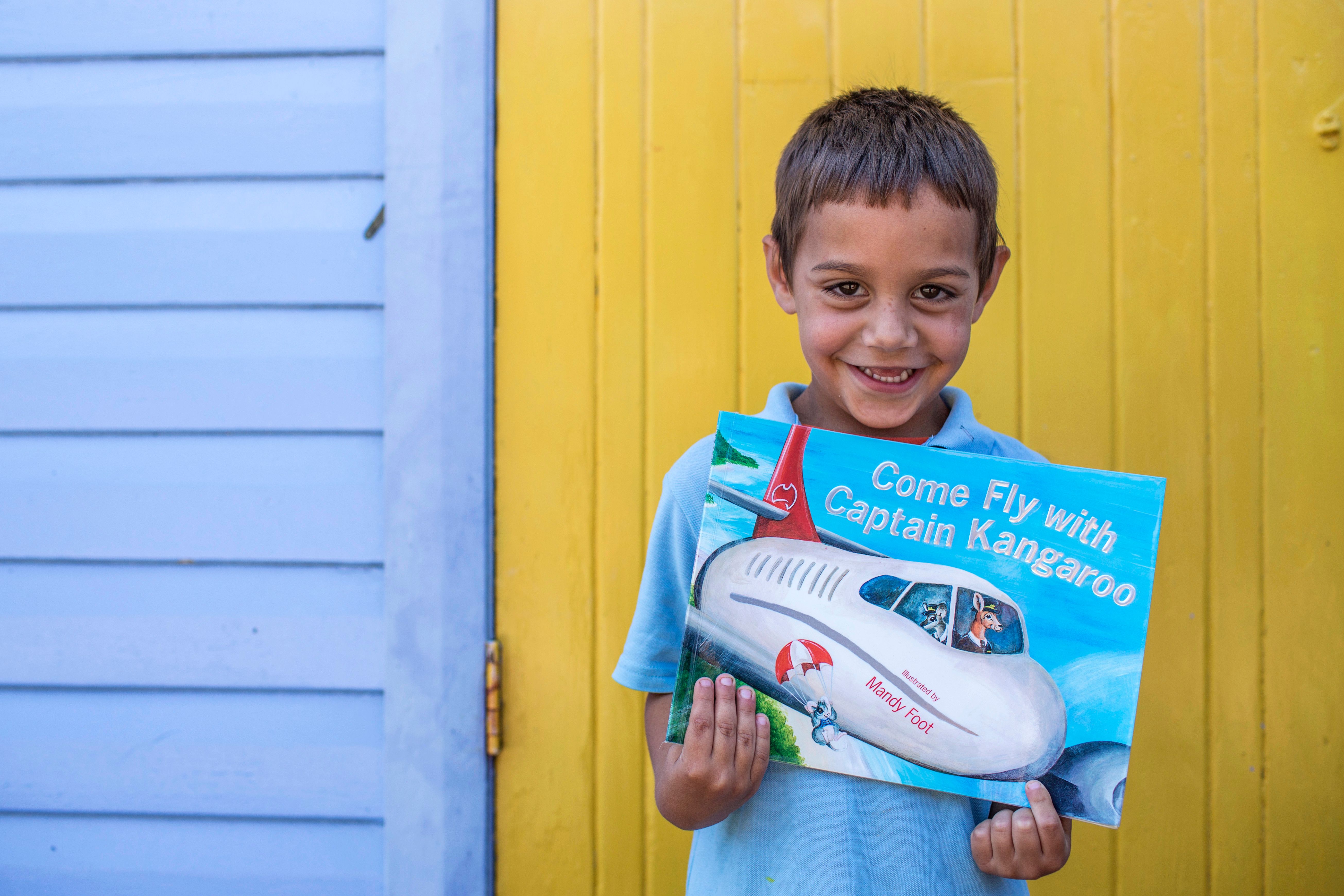 Child smiling and holding a book in front of a yellow door and a blue wall