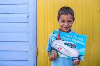 Child smiling and holding a book in front of a yellow door and a blue wall