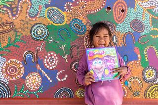 A child holding a book, laughing toward the camera in front of Indigenous artwork