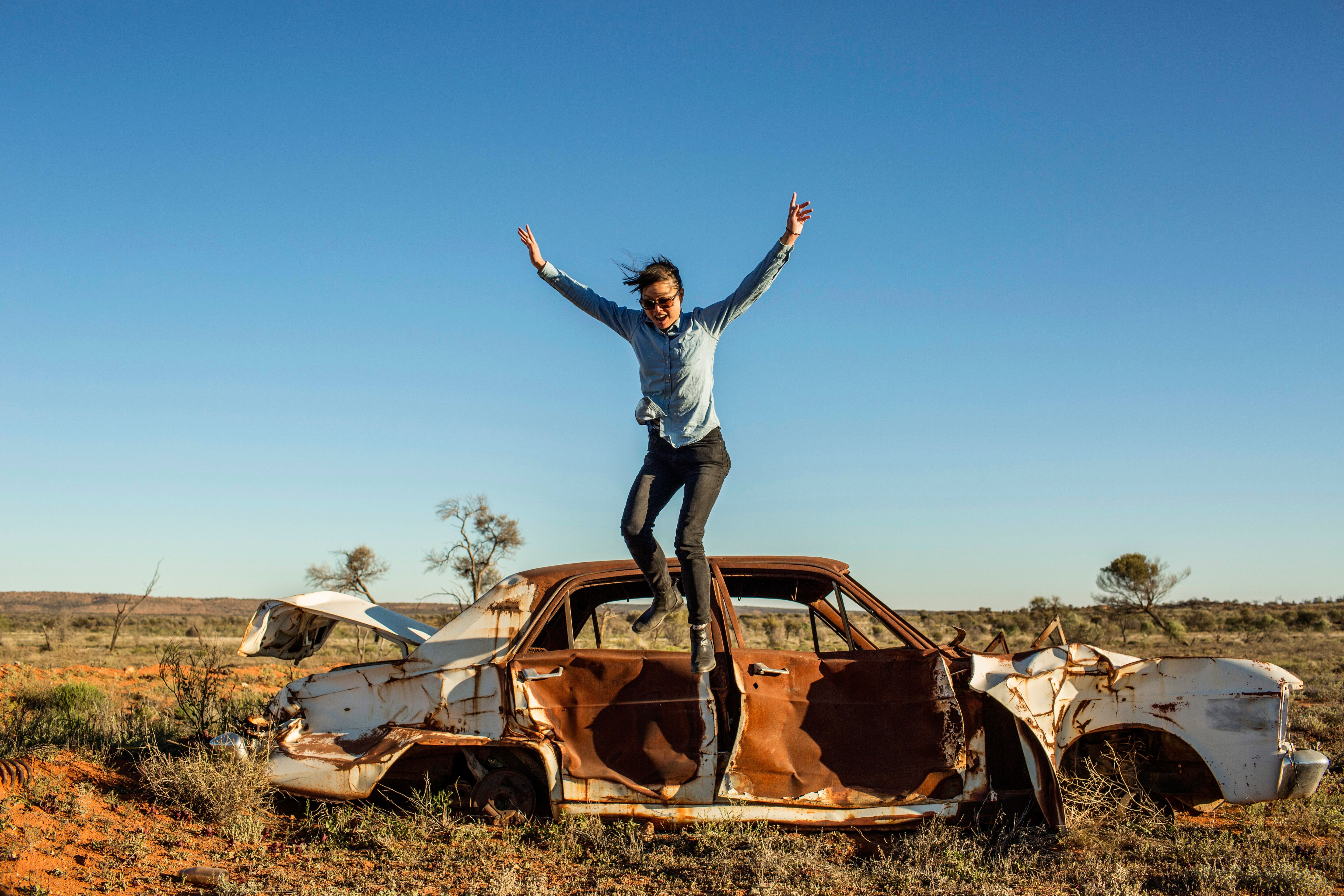 A woman mid-jump off the roof of an old car out in the bush