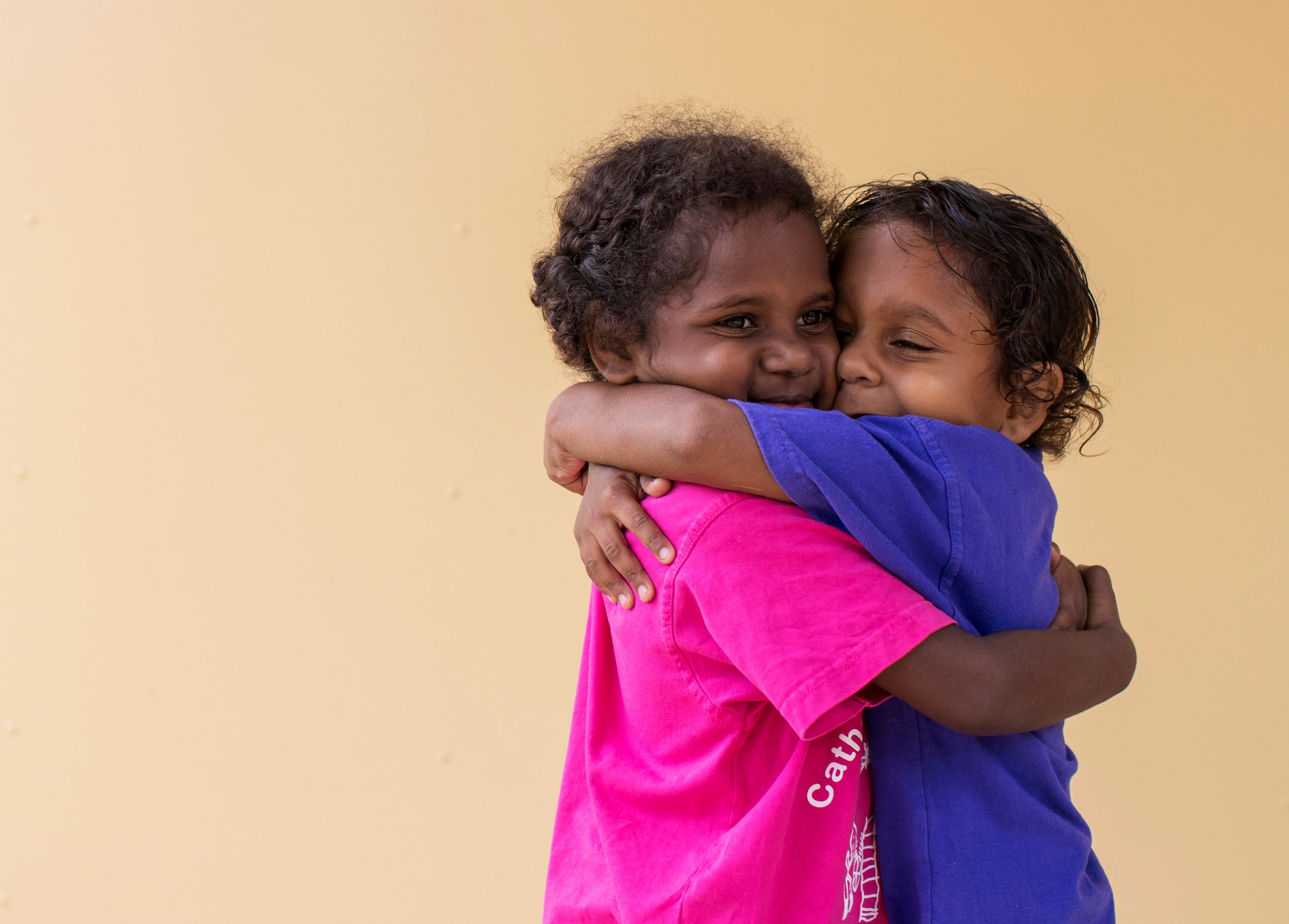 Two kids hugging each other in front of a plain background 