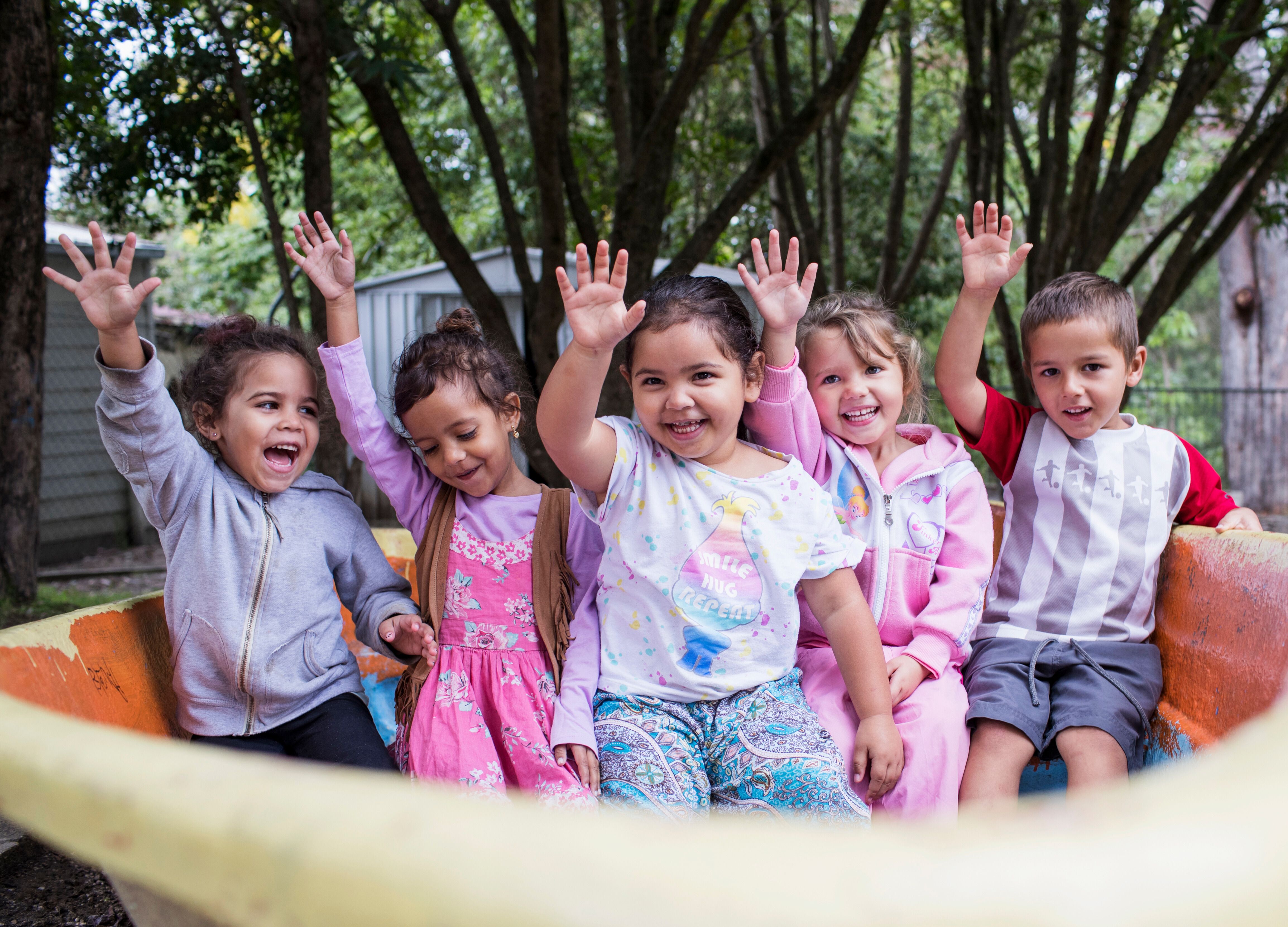 A group of children sitting on a playground outside smiling and holding their hands in the air 