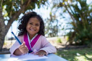 A child sitting at a table outside smiling toward the camera holding a pencil and paper