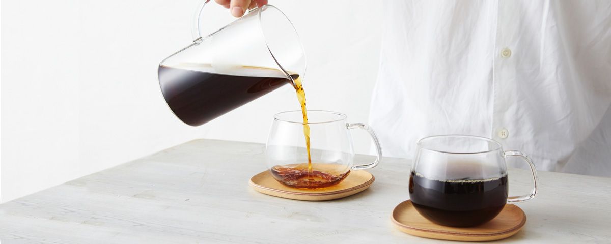 Pouring hot coffee from a glass carafe into a Kinto glass mug on a saucer.