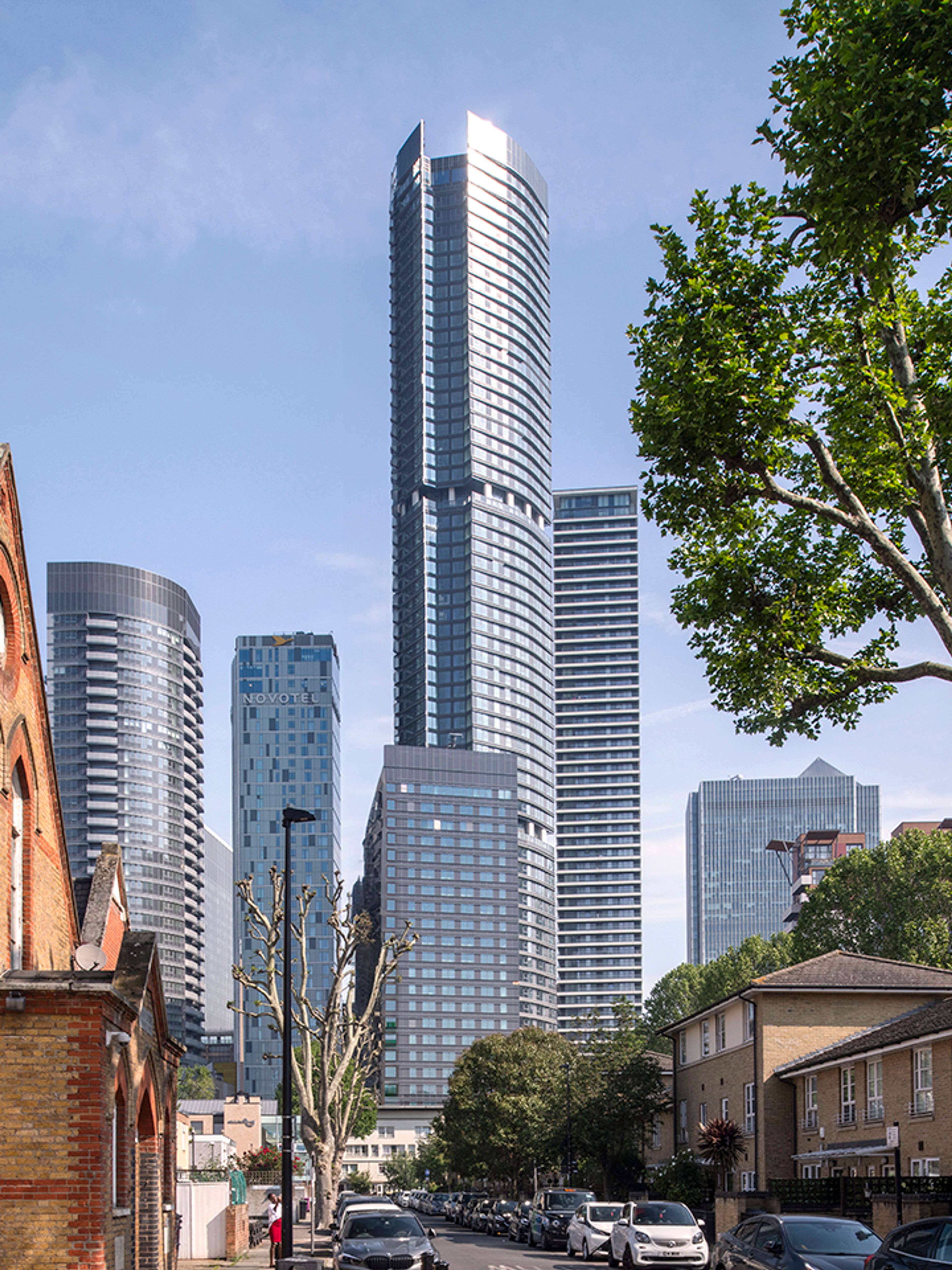 A tall and slender silver tower emerges from a similar coloured building cluster in Canary Wharf.