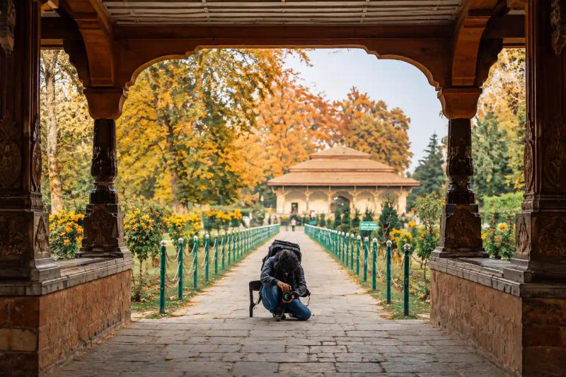 Photographer crouching on a stone path at Nishat Bagh Srinagar, framed by a carved wooden pavilion and autumn tree