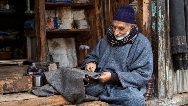 Elderly Kashmiri man in a woollen pheran stitching fabric, part of Kashmir winter food and culture