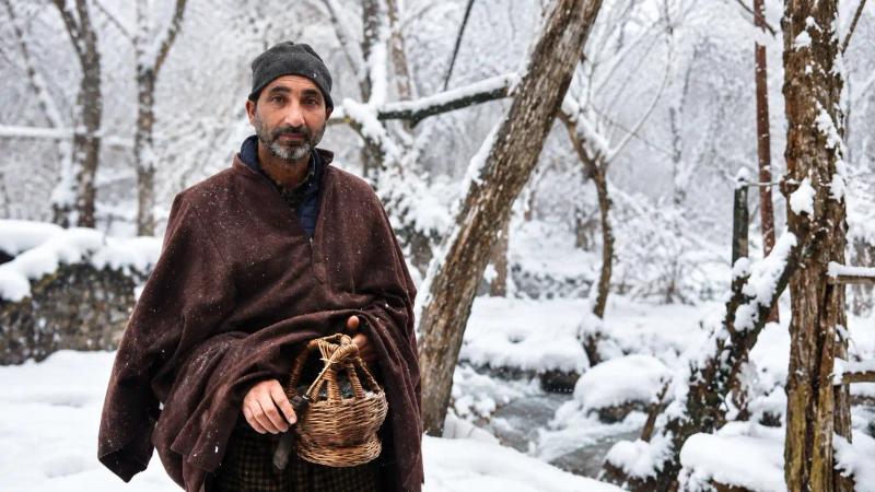 Kashmir winter food and culture showing a local man carrying a traditional wicker basket in a snow-covered forest during winter.