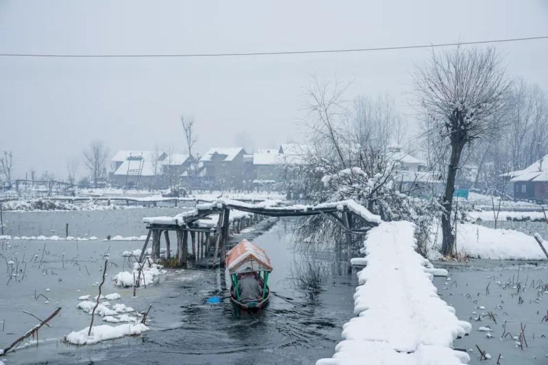 Chillai Kalan in Kashmir showing a traditional Kashmiri boat moving through partially frozen water surrounded by snow-covered houses.