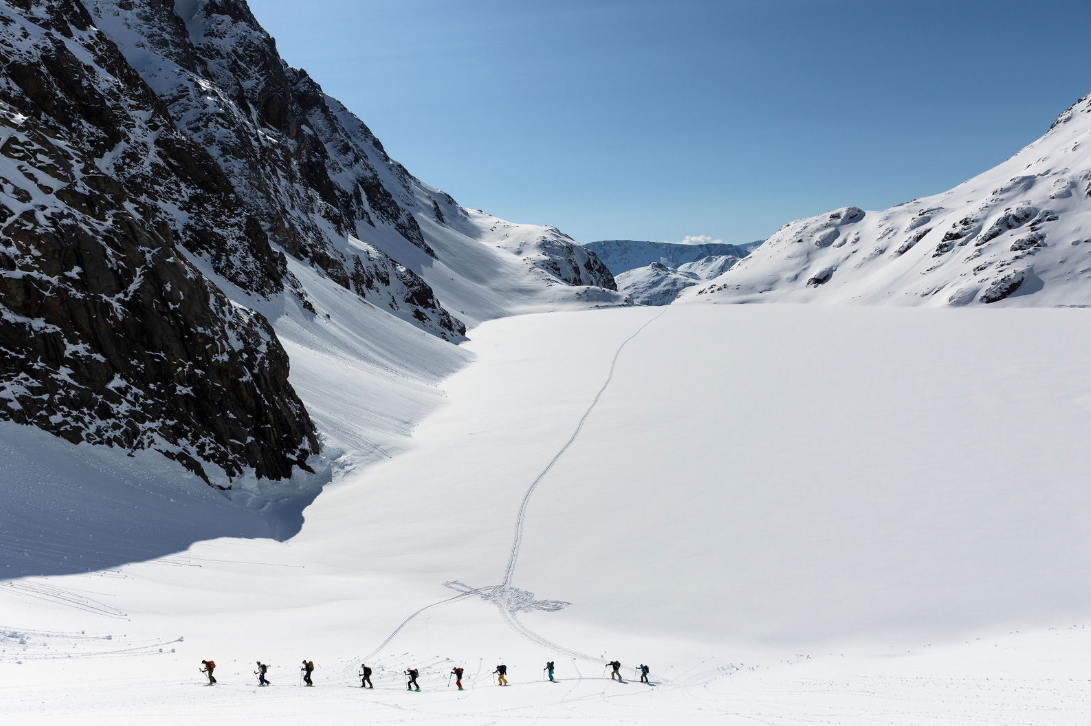 A line of skiers ascends a wide, snow-covered valley between steep, rocky mountain walls, following a single track across the vast white landscape under a clear blue sky.