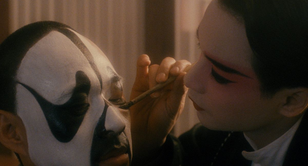 A performer in pink and white opera makeup uses a brush to finish the black and white face paint of another performer.