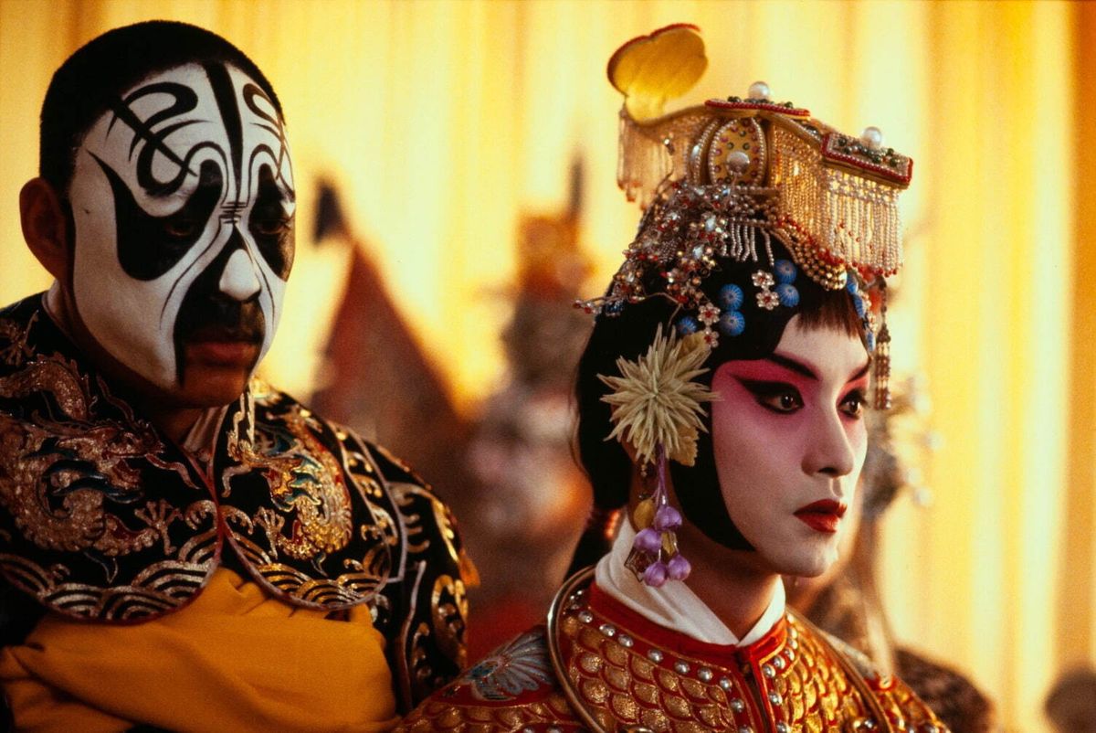 Two Beijing opera performers stand side by side in elaborate stage makeup and embroidered costumes against golden curtains.