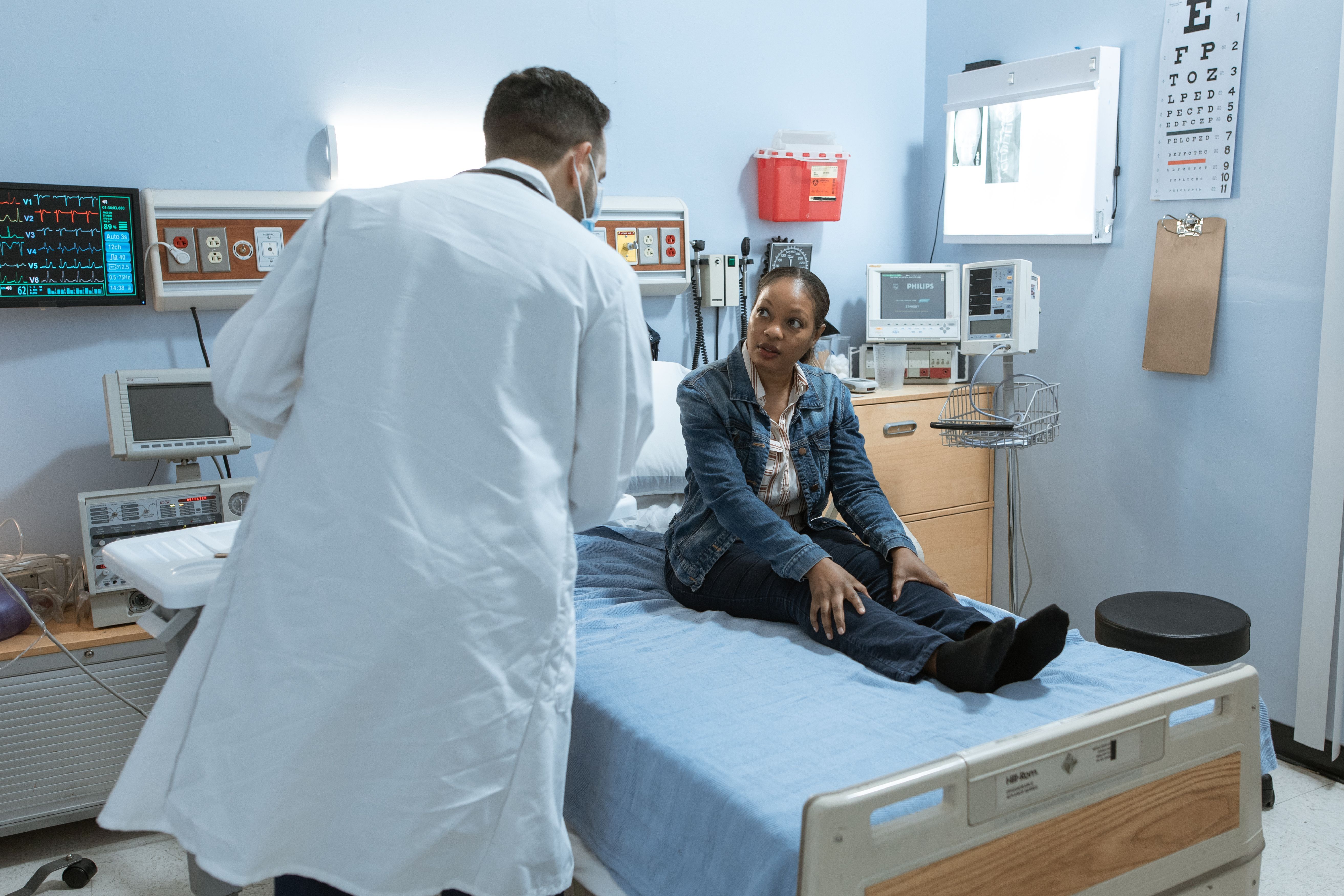 A doctor in a white coat consults with a female patient seated on an exam table in a medical examination room, with EKG monitors and medical equipment visible in the background.