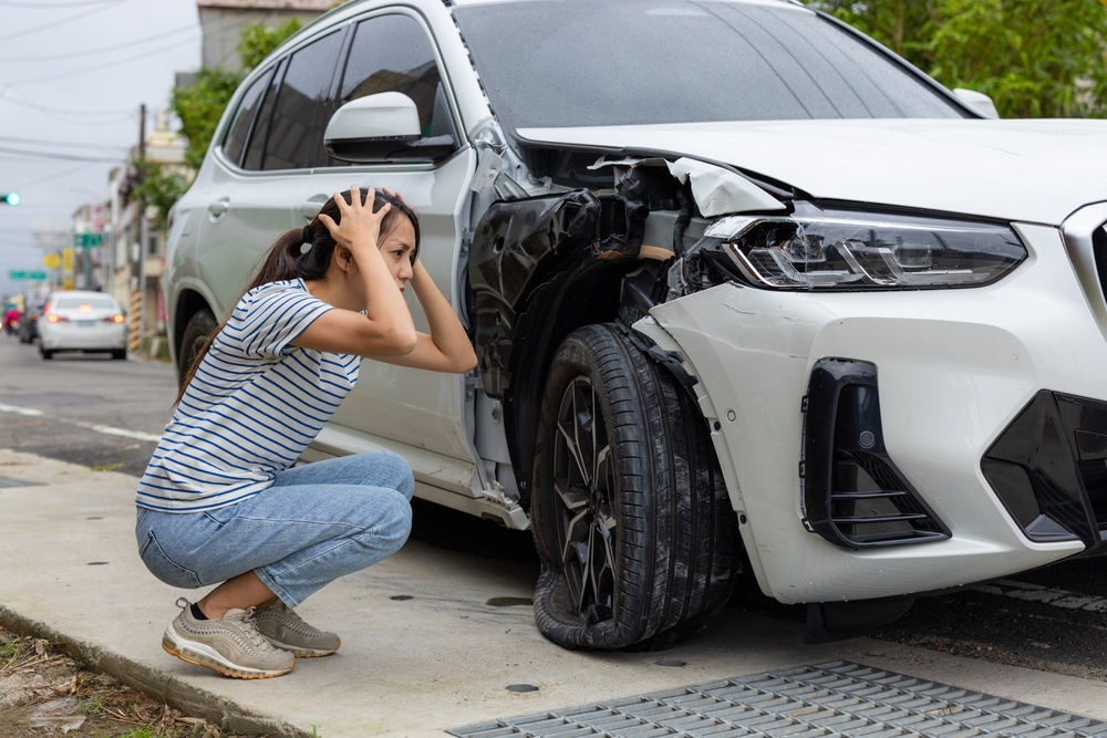 Image Woman crouching next to a damaged SUV after a car accident showing front-end impact and visible vehicle damage after a low-speed crash