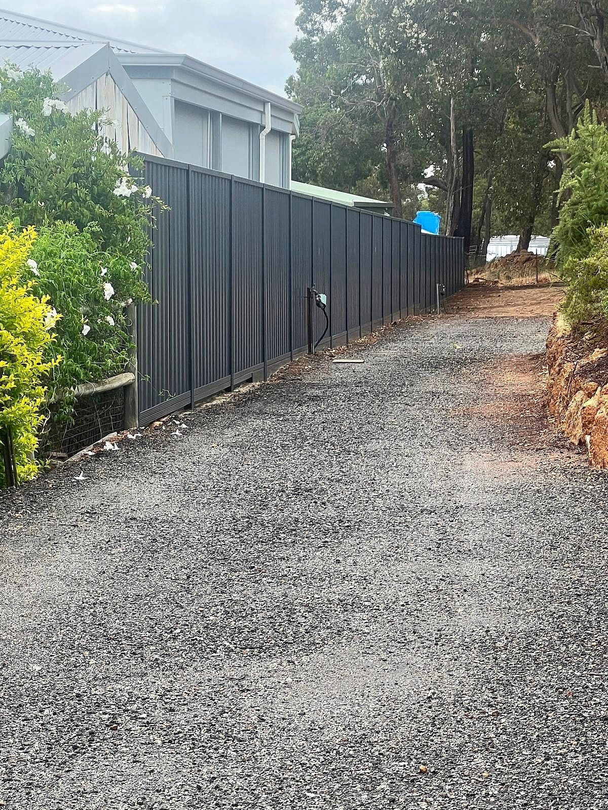 Dark charcoal Colorbond fence bordering a gravel driveway beside a country home surrounded by tall gum trees