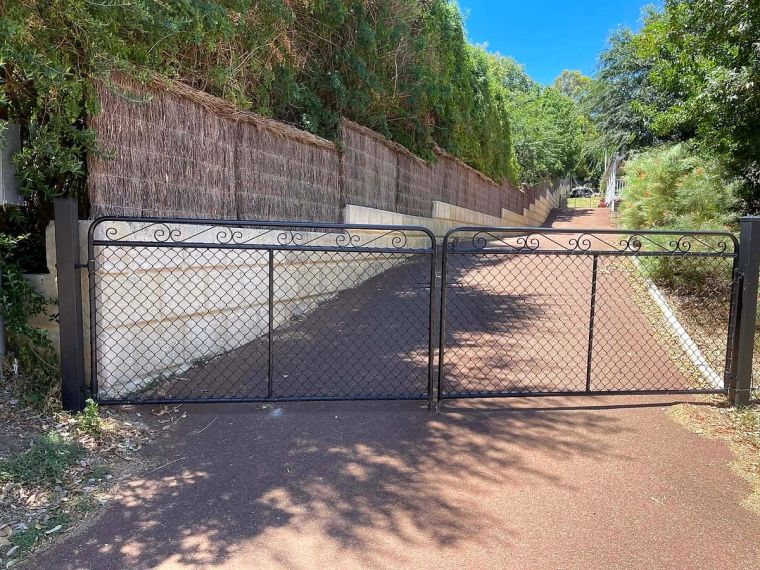 Natural brush fencing topped with greenery beside a black wrought-iron double gate leading to a Perth Hills driveway
