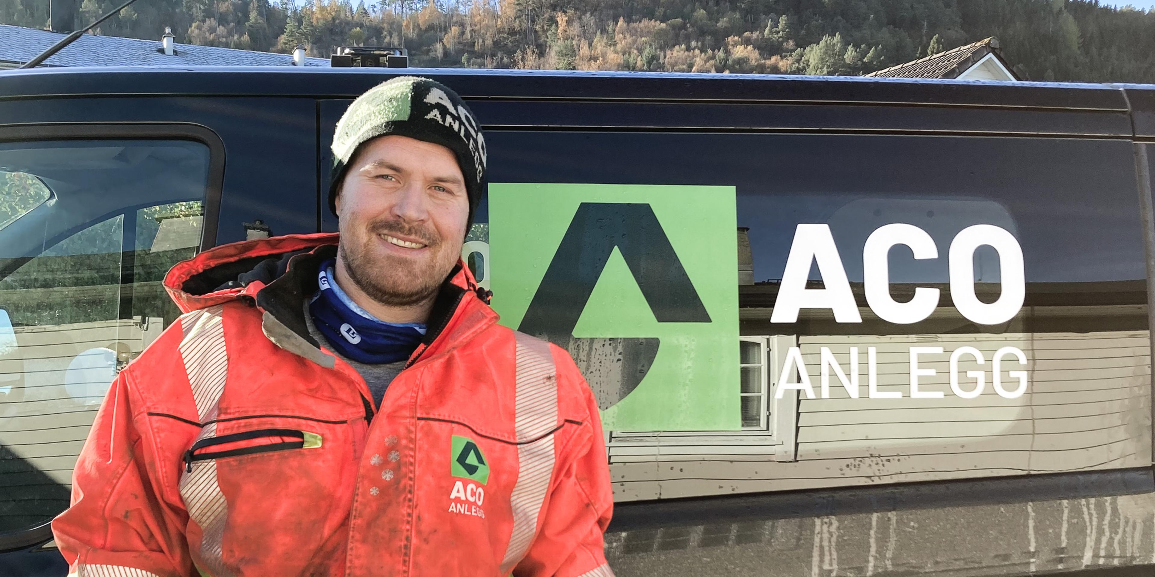 En mann i en oransje jakke foran en bil Aco Anlegg
