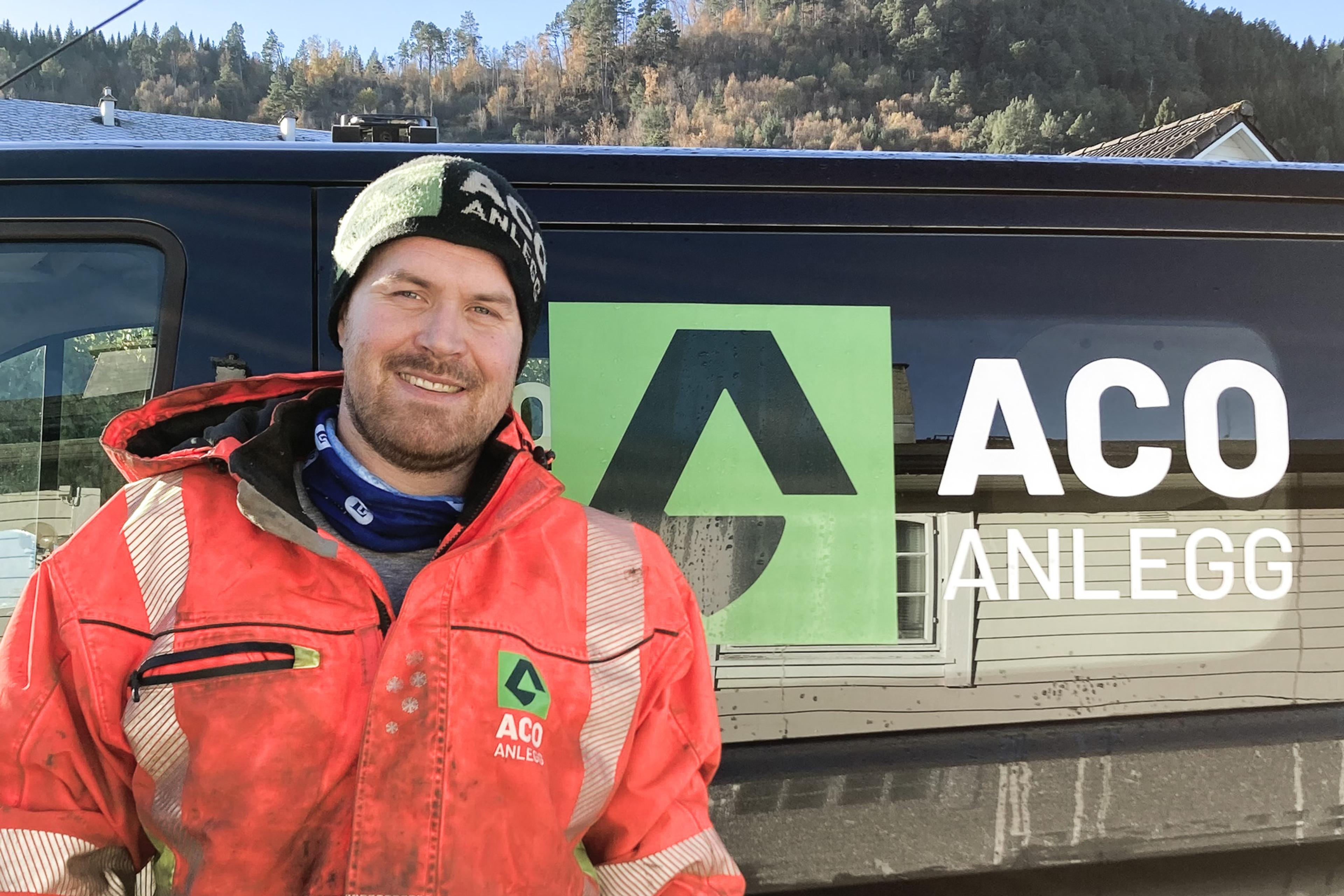 En mann i en oransje jakke foran en bil Aco Anlegg