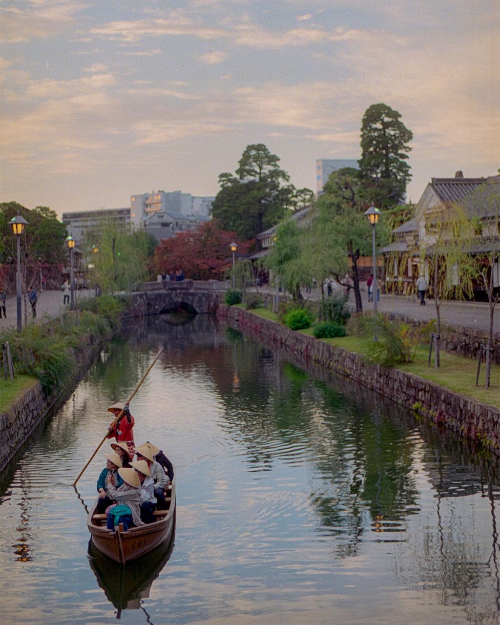 Traditional boat carrying visitors along the historic canal of Kurashiki, Japan, lined with white-walled storehouses and willow trees at dusk.