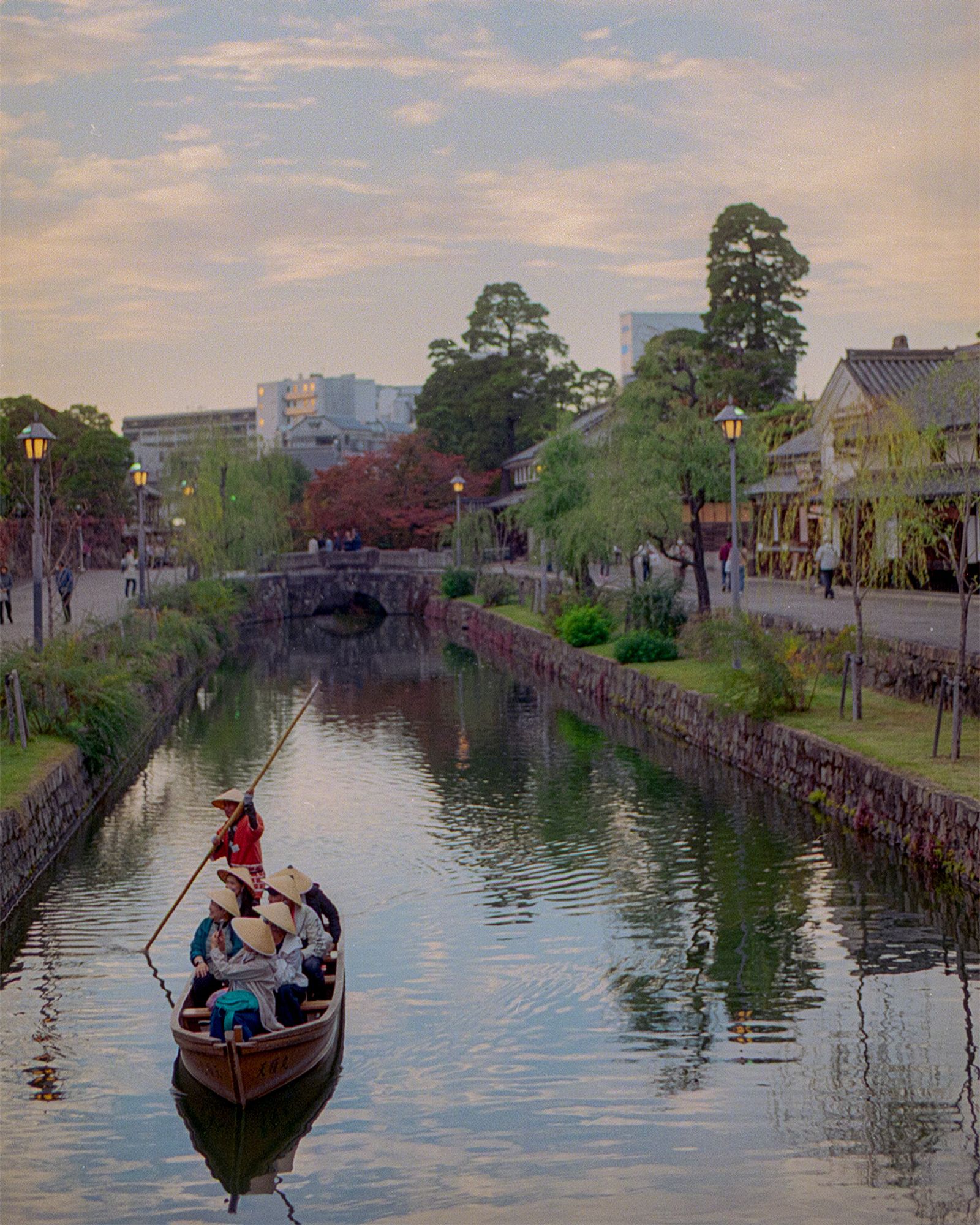 Traditional boat carrying visitors along the historic canal of Kurashiki, Japan, lined with white-walled storehouses and willow trees at dusk.