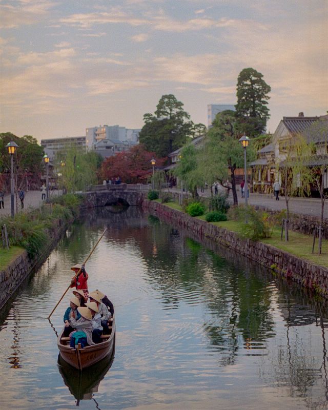 Traditional boat carrying visitors along the historic canal of Kurashiki, Japan, lined with white-walled storehouses and willow trees at dusk.