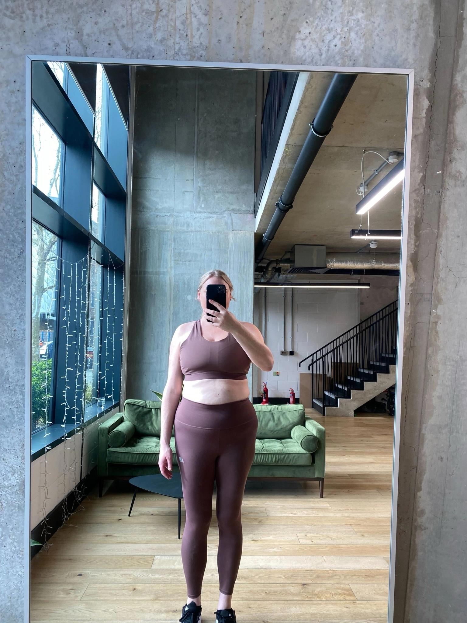 Full-body mirror photo. Person is wearing close-fitting athletic clothing, with body shape clearly visible. Face is partially obscured by phone.