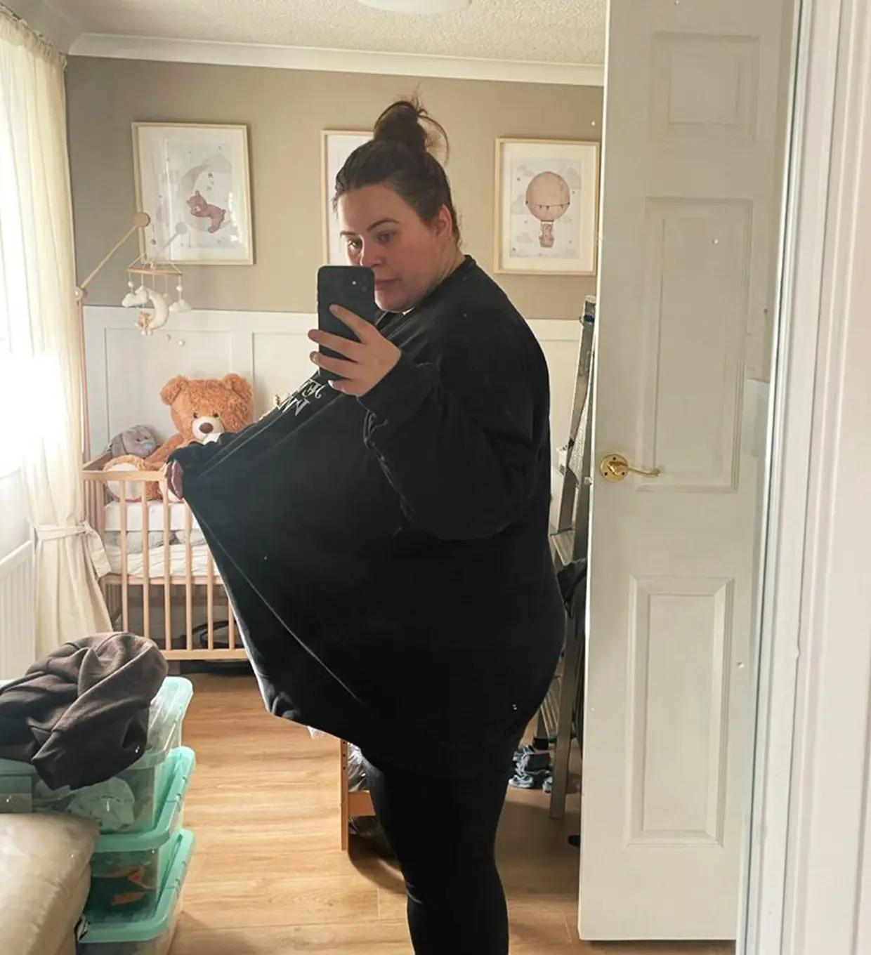 Kelsey taking a mirror selfie in a bedroom, holding out an oversized black top to highlight weight loss achieved with Mounjaro as part of the Voy programme.
