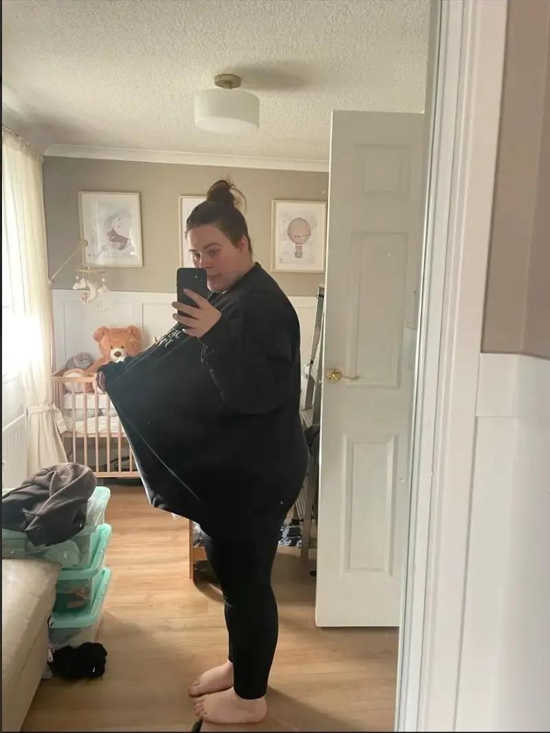 Kelsey taking a mirror selfie in a bedroom, holding out an oversized black top to highlight weight loss achieved with Mounjaro as part of the Voy programme.