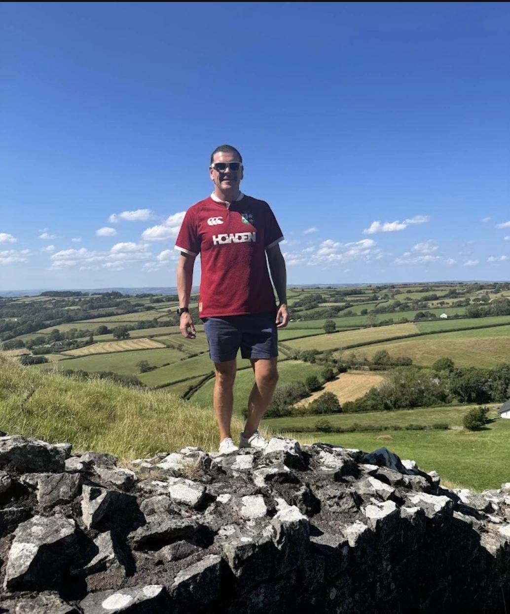 Man standing on a stone wall wearing a red rugby shirt with countryside and blue sky in the background