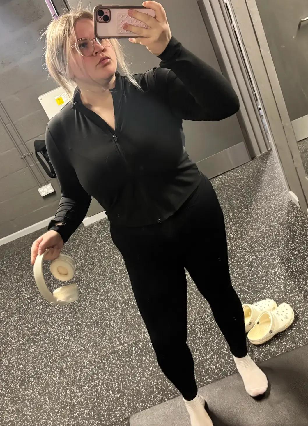Shania taking a mirror selfie at the gym, wearing black activewear and holding headphones, showing her weight loss progress as part of her journey with Mounjaro and the Voy programme.