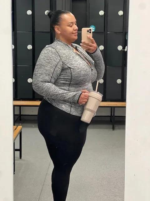 Hollie in a gym locker room smiling and holding a protein shake, wearing a grey zip-up top and black leggings, proudly capturing a moment in her weight loss journey.