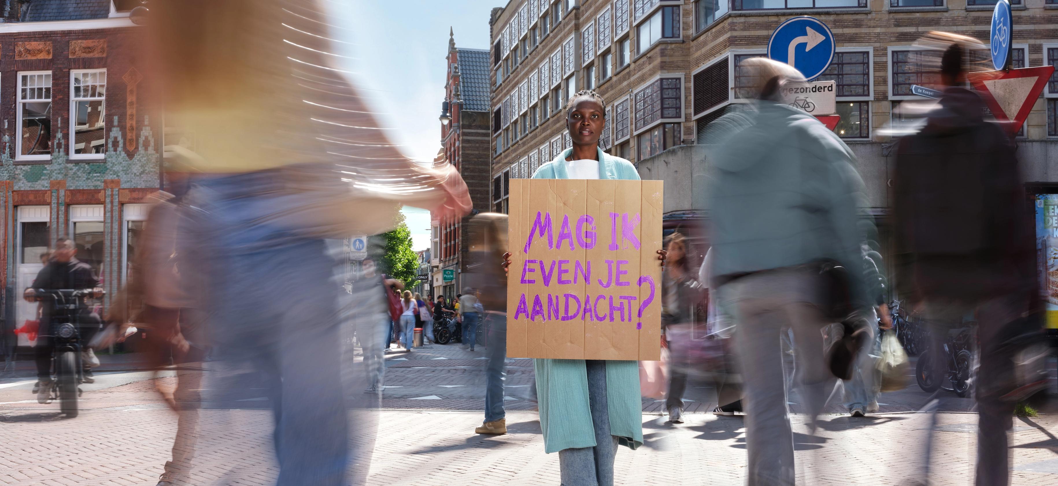Vrouw houdt een bord vast met "mag ik even je aandacht" in het midden van een drukke straat.