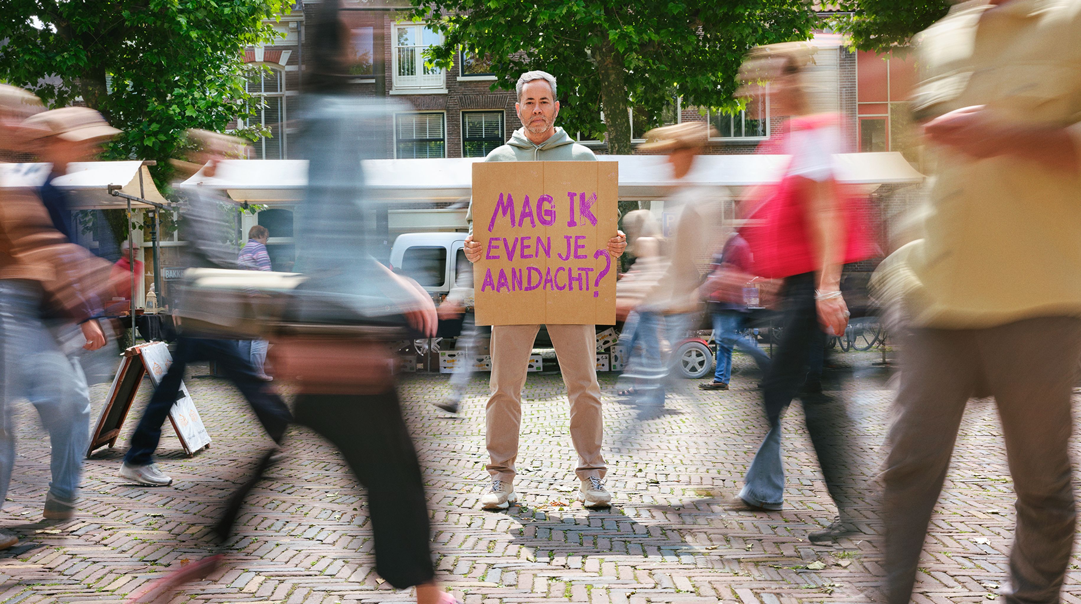 Key visual met een man met een bord waarop "mag ik even je aandacht" staat geschreven, in het midden van een drukke winkelstraat.