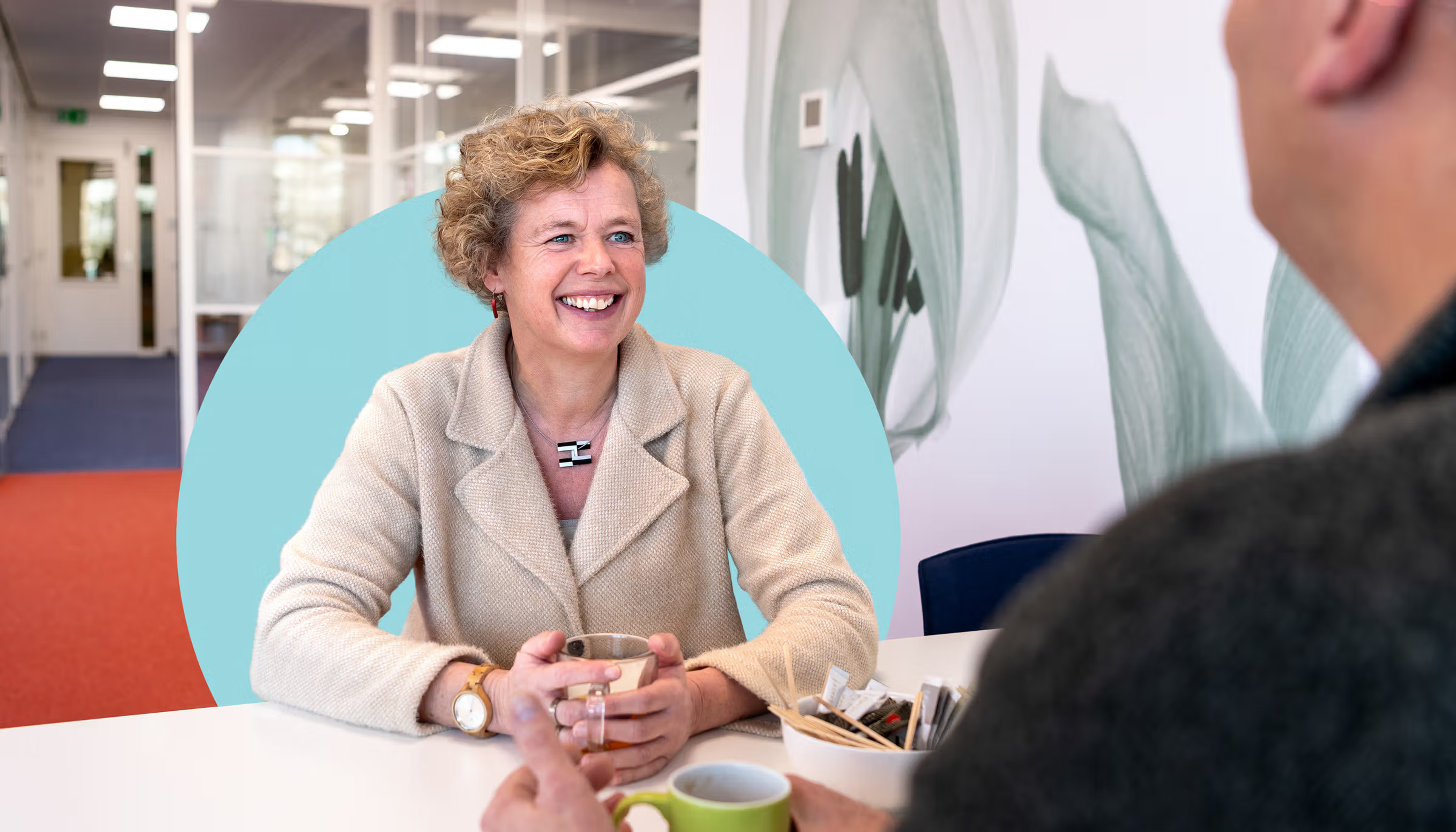 Vrouw in rode blazer en zwarte top in gesprek, omkaderd door een turquoise cirkel, in een lichte binnenruimte.