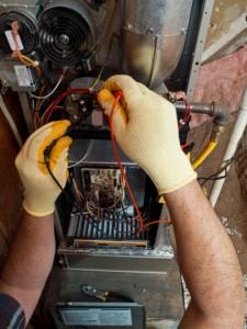Gloved hands testing the wiring inside an open furnace with a multimeter