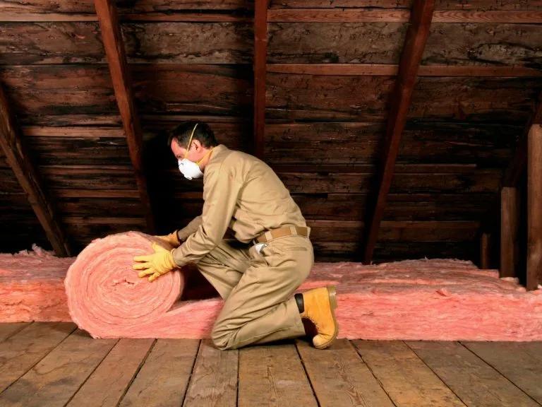 Worker in a mask unrolling pink fiberglass insulation in an attic