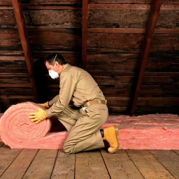 Worker in a mask unrolling pink fiberglass insulation in an attic