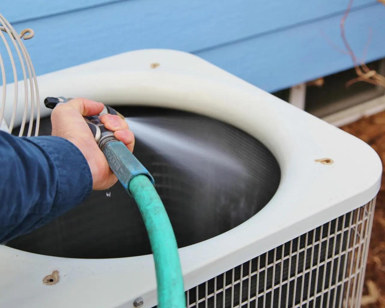 Person rinsing the top of an outdoor air conditioning unit with a garden hose