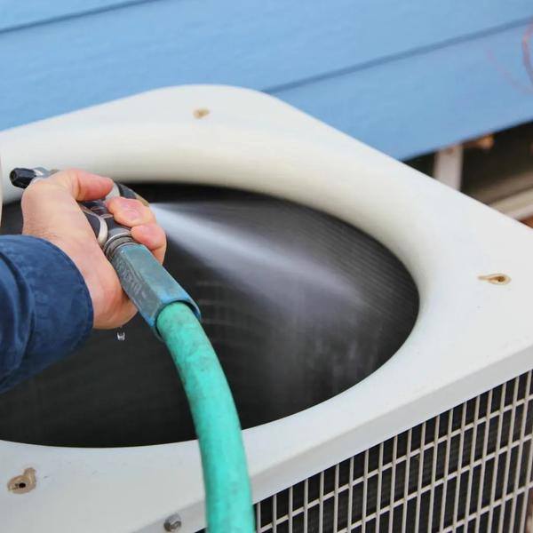 Person rinsing the top of an outdoor air conditioning unit with a garden hose
