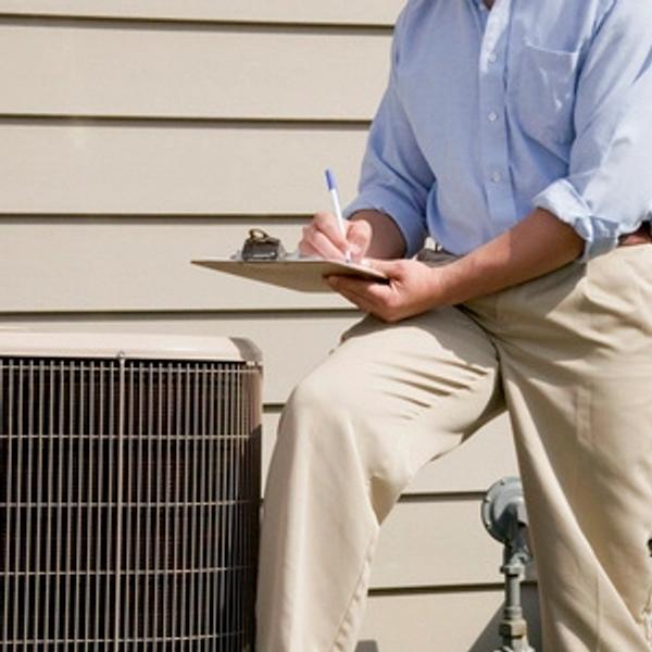 Inspector with a clipboard sitting on an outdoor air conditioning unit taking notes