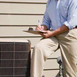 Inspector with a clipboard sitting on an outdoor air conditioning unit taking notes