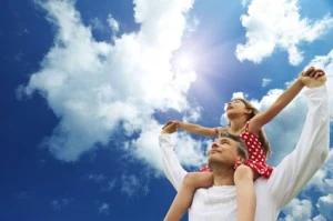 Child riding on a parent's shoulders with arms outstretched under a bright blue sky