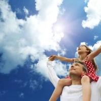 Child riding on a parent's shoulders with arms outstretched under a bright blue sky