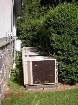 Outdoor air conditioning unit crowded by overgrown shrubs beside a house