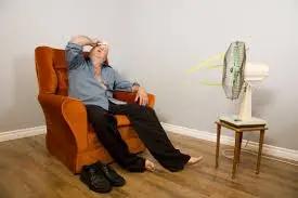 Man slumped in an armchair in front of a pedestal fan, wiping his forehead