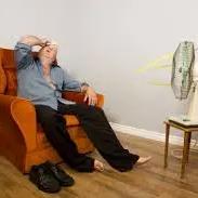 Man slumped in an armchair in front of a pedestal fan, wiping his forehead