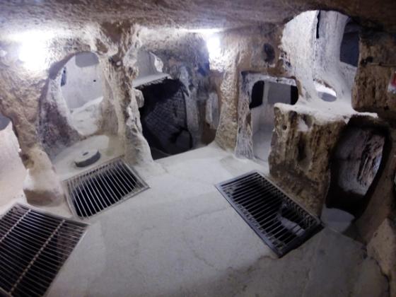 A carved underground chamber at Kaymaklı Underground City, Cappadocia, showing rock-hewn passageways, a circular stone door, and modern lighting and safety grates.