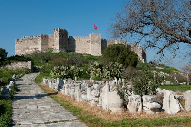 The crenellated stone walls and towers of Selçuk Castle on Ayasuluk Hill, flying a Turkish flag, approached past ancient marble column fragments.